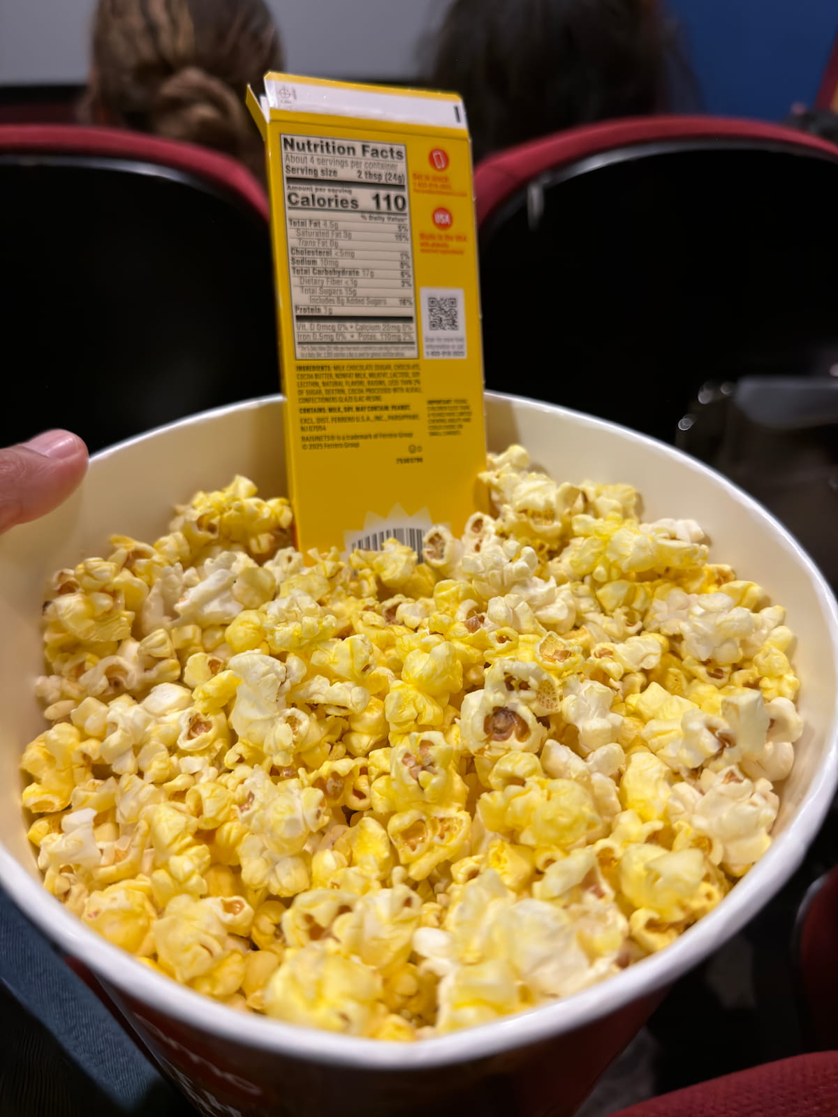 A bucket of movie theater popcorn with a box of Raisinets stuck in the middle
